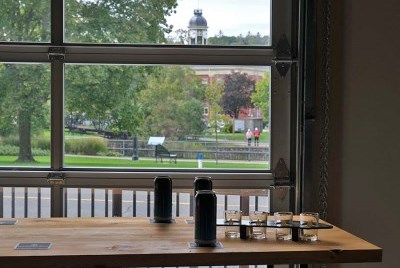 A table set with beer cans and flights in front of a large glass garage door, with a view of a park and a town hall.