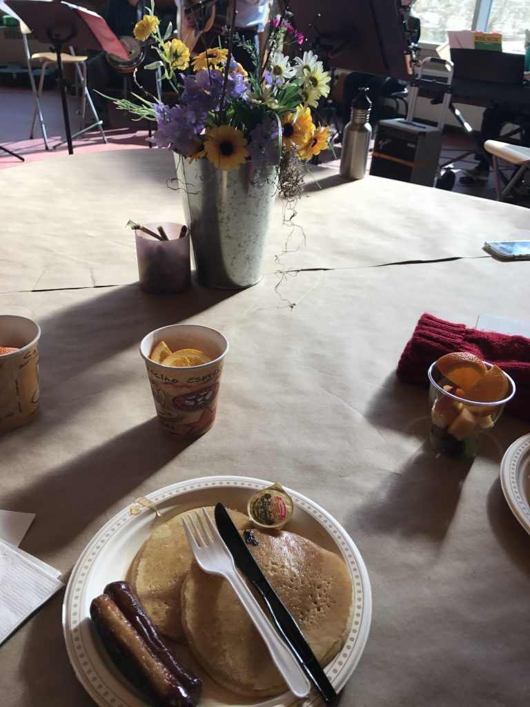 A foam plate in a white table with a pancake and two breakfast sausages. There are some yellow and purple flowers also on the table while a band plays in the background.