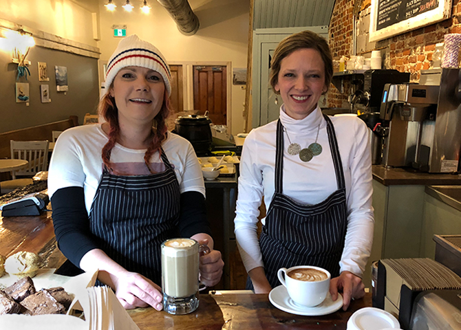 Servers Erin and Ginn smiling and holing lattes behind the counter at Happenstance Coffee Public