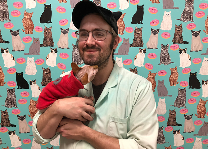 Owner of Toe Beans Cafe holding cat in front of wallpaper featuring cats and donuts 