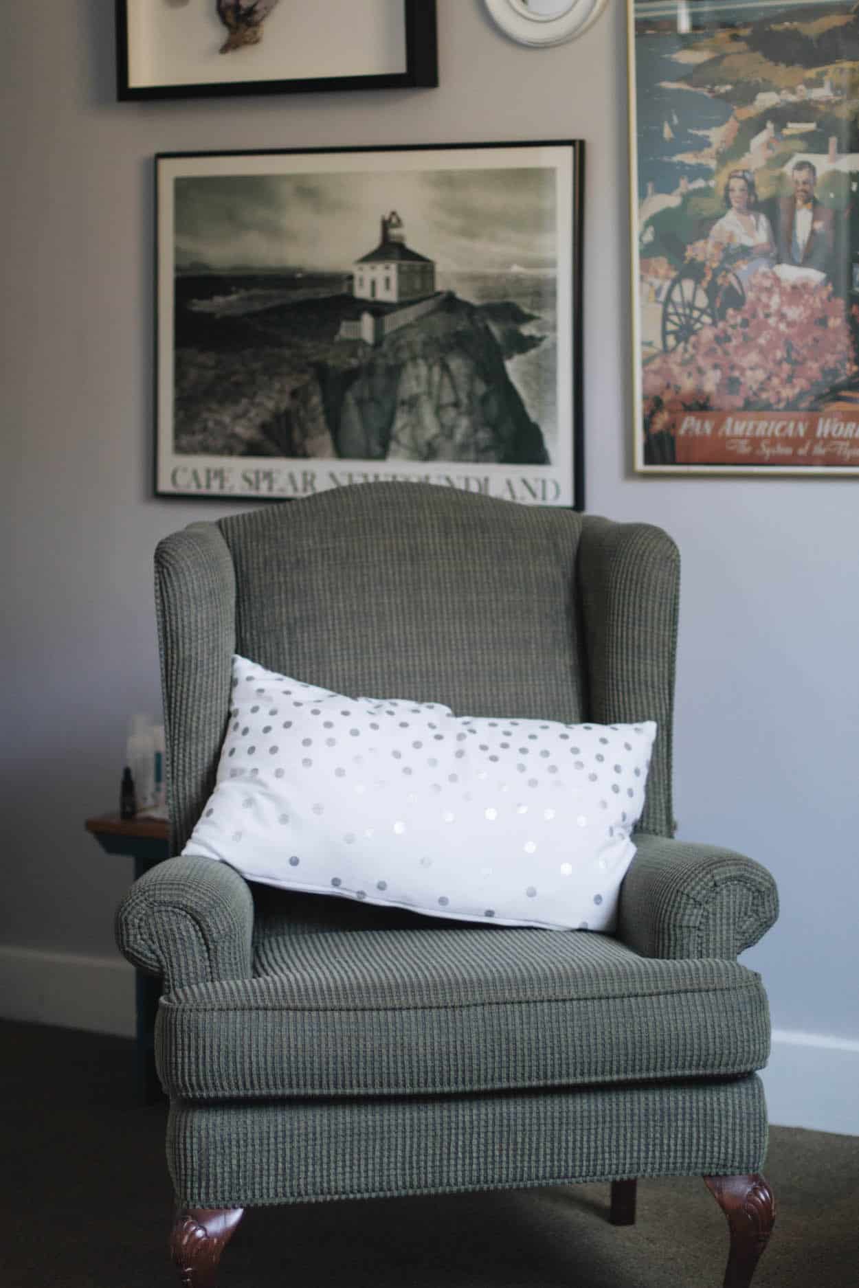 A photo of a grey corduroy chair and a white pillow with silver polka-dots in front of old posters in a bedroom at the Hotel Carlyle