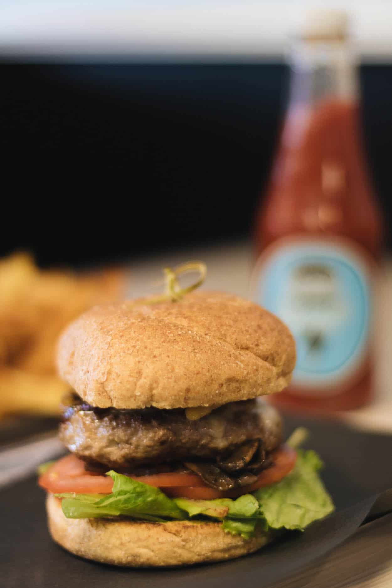 A photo of the Zeus Burger with lettuce, tomato, carmalized onions, sauteed mushrooms, and cheddar cheese in front of a bottle of ketchup