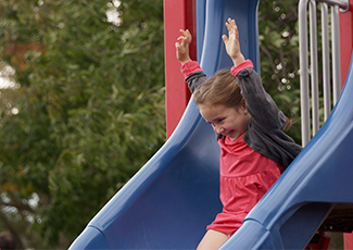 A little girl going down a slide, with leafy trees in the background.